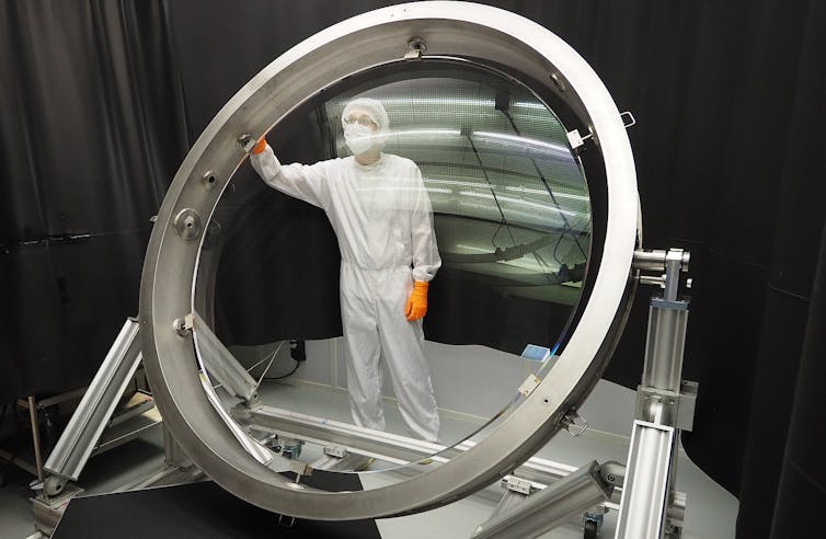 A large round, convex glass lens mounted on a metal stand, with a technician wearing scrubs looking at it.