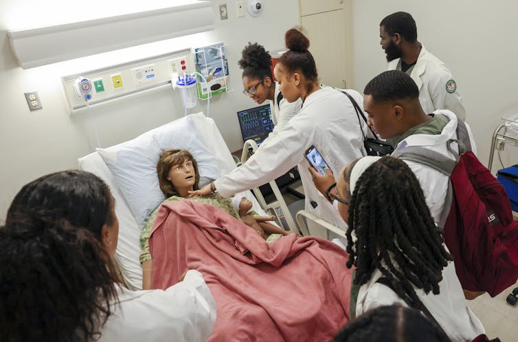 A group of young people wear white jackets and stand around a dummy dressed with a pink blanket over it in a hospital bed.